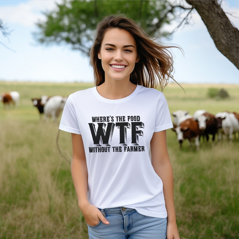 K&ouml;nnte beinhalten: Wei&szlig;es T-Shirt mit dem Text "WHERE'S THE FOOD WTF WITHOUT THE FARMER" in Schwarz. Die Person tr&auml;gt das Shirt auf einer Wiese mit K&uuml;hen im Hintergrund. Die Person l&auml;chelt.
