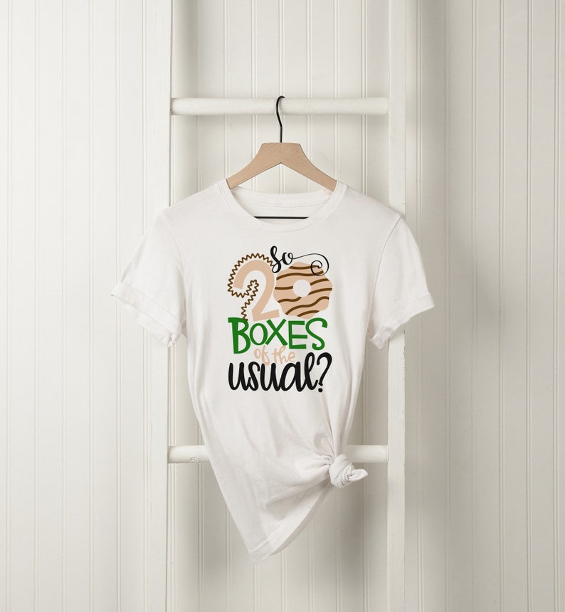 May include: White t-shirt with a graphic design featuring the text "So 20 Boxes of the usual?" in green and black. The design includes a stylized number 2 and a donut illustration in tan and brown.