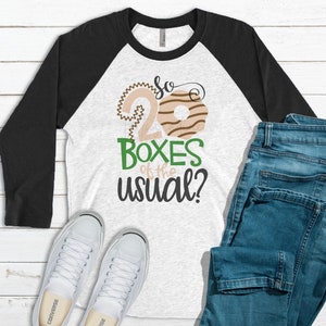 May include: A white and gray raglan shirt with black sleeves, featuring the text "So 20 Boxes of the usual?" in a playful font. The design includes donut graphics. The image also shows blue jeans and white sneakers.