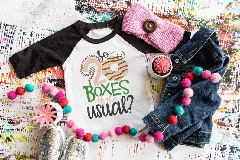 May include: A white and black raglan t-shirt with the text "So 20 Boxes of the usual?" is displayed with a denim jacket, pink headband, and colorful pom-pom garland. A small succulent in a white pot and silver shoes are also present.