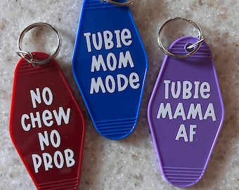 May include: Three colorful keychains with silver key rings. One is red with the text "NO CHEW NO PROB", one is blue with "TUBIE MOM MODE", and the last is purple with "TUBIE MAMA AF". The keychains are in a retro motel style.