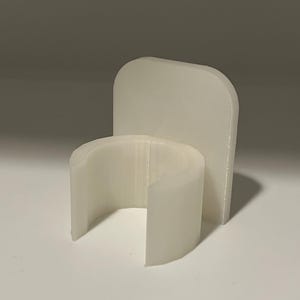 May include: A white, 3D-printed plastic holder featuring a semi-circular, open-front clip attached to a flat, rounded rectangular backplate. The object has a smooth, matte finish and a uniform, opaque white color throughout its structure.