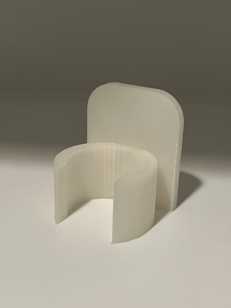 May include: A white, 3D-printed plastic holder featuring a semi-circular, open-front clip attached to a flat, rounded rectangular backplate. The object has a smooth, matte finish and a uniform, opaque white color throughout its structure.