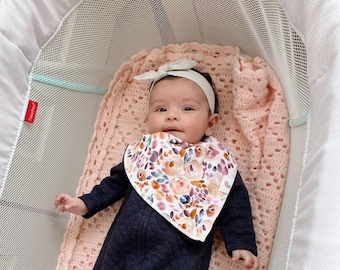 Può includere: Un bambino che indossa un pigiama blu scuro e un bavaglino floreale giace in un lettino rosa e bianco. Il bambino indossa una fascia bianca con un fiocco.