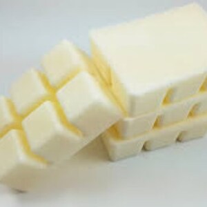 May include: Cream-colored wax melt cubes arranged in a stack and a row. The cubes are rectangular with rounded edges, suggesting a smooth texture. The wax melts are a light, off-white color.