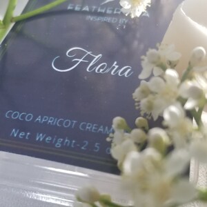 May include: A close-up of a product package with the word "Flora" in elegant script. The package is surrounded by white flowers and greenery. The text "Coco Apricot Cream" is visible.