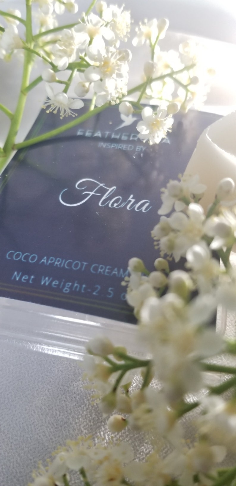 May include: A close-up of a product package with the word "Flora" in elegant script. The package is surrounded by white flowers and greenery. The text "Coco Apricot Cream" is visible.