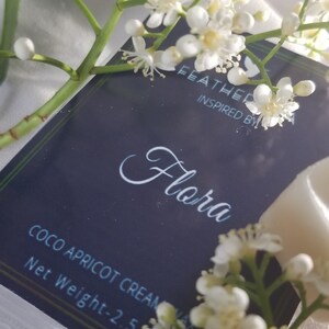 May include: A close-up of a product package with the word "Flora" in cursive script. The package is dark blue with white text and is surrounded by white flowers. The net weight is 2.5 oz.