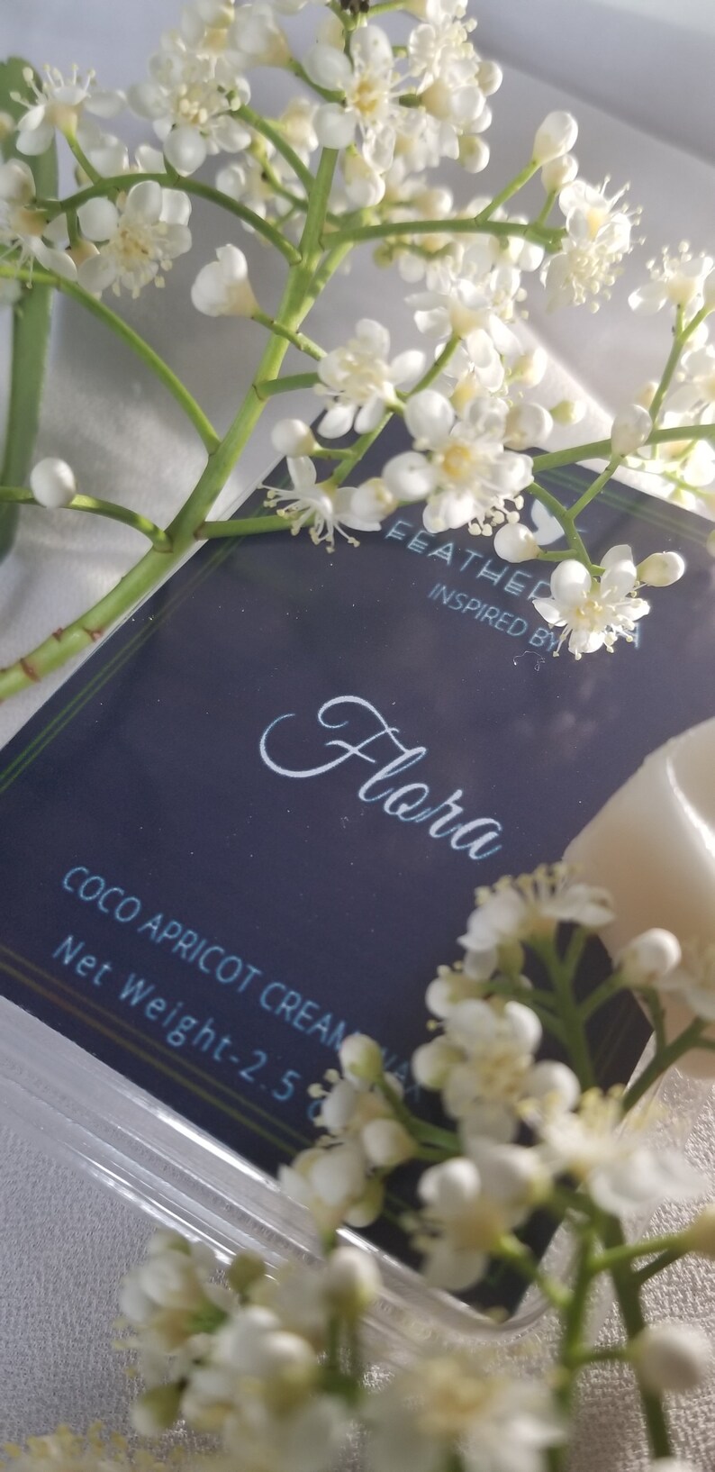 May include: A close-up of a product package with the word "Flora" in cursive script. The package is dark blue with white text and is surrounded by white flowers. The net weight is 2.5 oz.