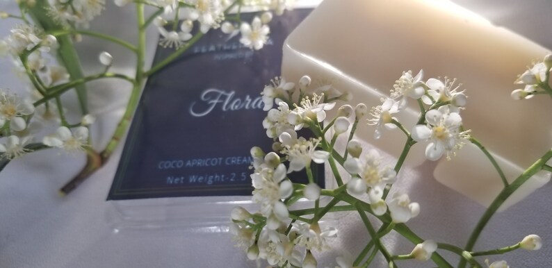 May include: A bar of light-colored soap and a package of Coco Apricot Cream are surrounded by white flowers. The package has the word "Flora" in cursive and the text "Net Weight-2.5 oz."