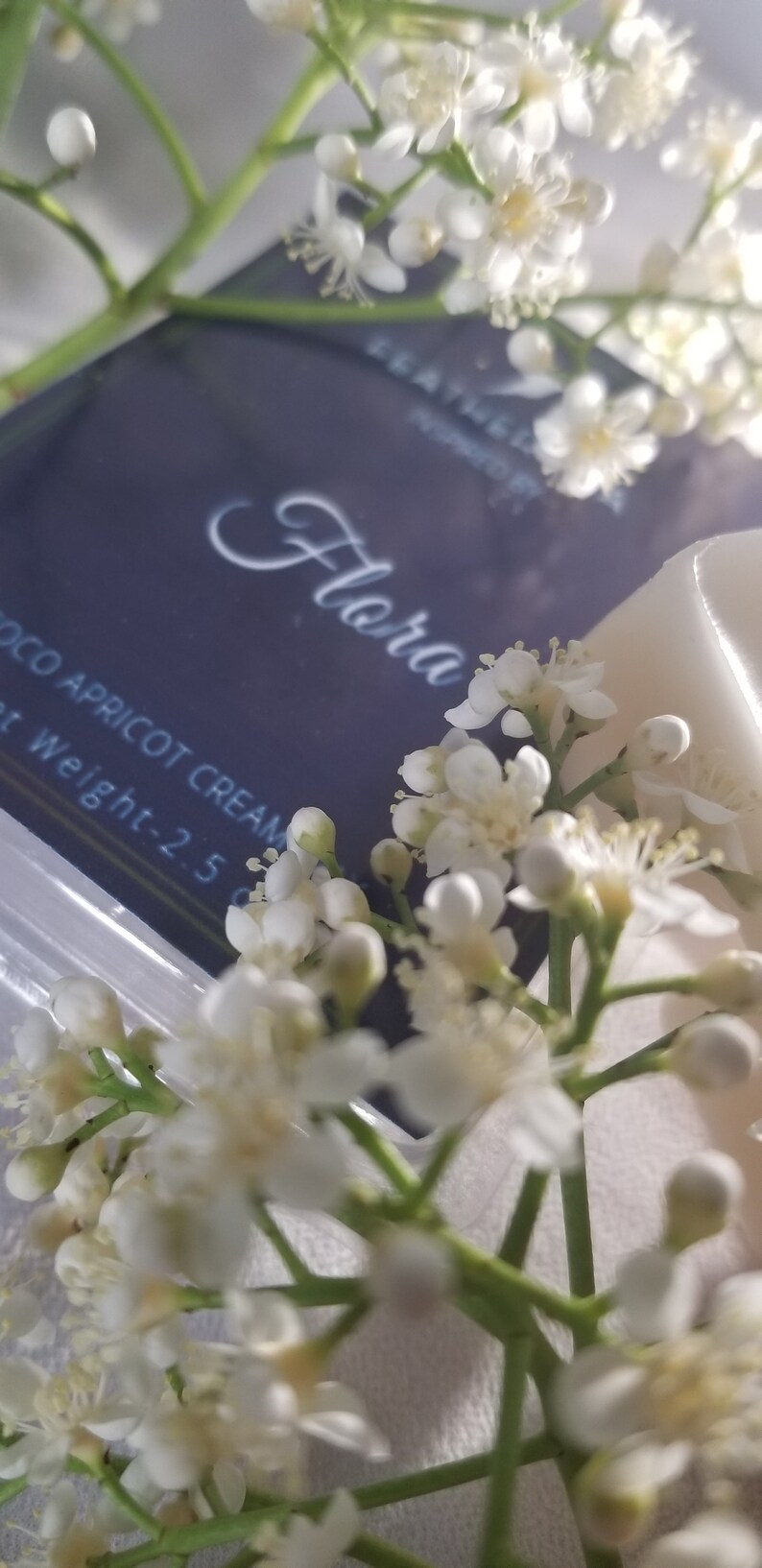May include: A close-up shot of a dark blue product box with the word "Flora" in elegant script. Delicate white flowers and buds frame the box, with the text "COCO APRICOT CREAM" visible. The net weight is 2.5 oz.
