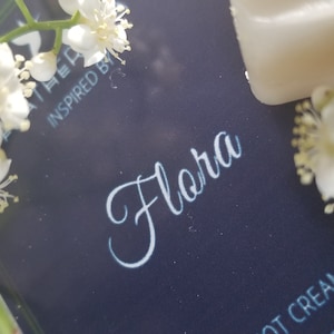 May include: A close-up of a dark blue product label with the word "Flora" in elegant white script. The label is surrounded by delicate white flowers and greenery. The text "INSPIRED BY" is visible. The image suggests a floral-themed product.