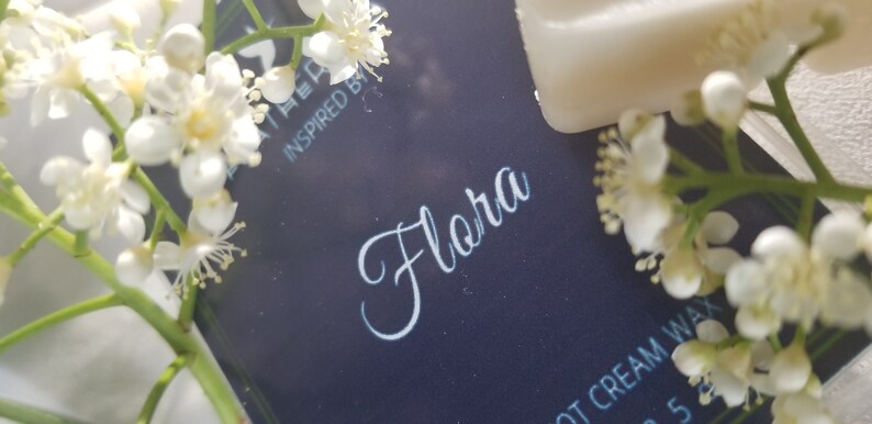May include: A close-up of a dark blue product label with the word "Flora" in elegant white script. The label is surrounded by delicate white flowers and greenery. The text "INSPIRED BY" is visible. The image suggests a floral-themed product.