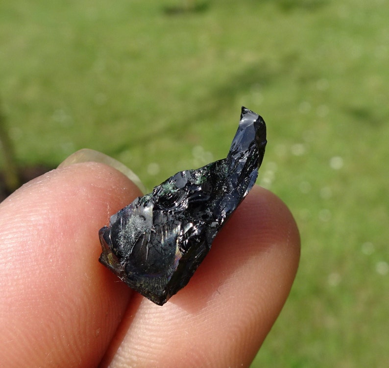 Puede incluir: Un cristal de obsidiana negro con una punta afilada y matices iridiscentes verdes y azules.