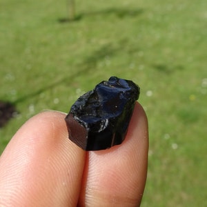 Puede incluir: Una roca de obsidiana negra con una superficie rugosa e irregular. La roca es del tama&ntilde;o de una punta de dedo.