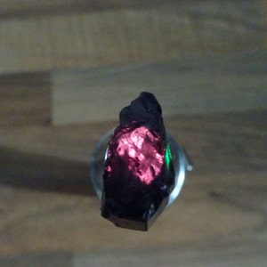 Puede incluir: Una piedra preciosa negra, rugosa y oscura con un brillo rojo iridiscente. La piedra preciosa est&aacute; montada en un soporte plateado.