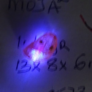 Può includere: Una pietra preziosa rosa con un bagliore viola chiaro sotto una luce UV. La pietra misura circa 13 x 8 mm.