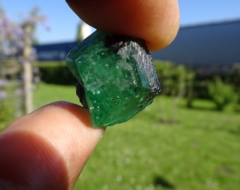 May include: A large, dark green emerald crystal with a rough, faceted surface. The crystal is held in a hand, with a blurred background of green grass and a blue sky.