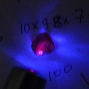 Può includere: Primo piano di una pietra preziosa rossa sotto una luce UV. La pietra preziosa è illuminata da un colore rosa brillante. Il testo "10 x g. 8 x 7" è visibile sullo sfondo.