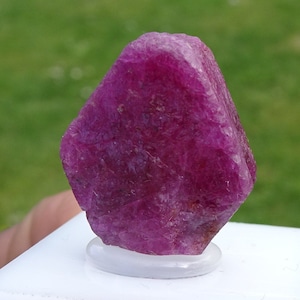 以下が含まれることがあります： 自然な粗い仕上げが施された濃いピンク色のルビーの宝石。ルビーは白いスタンドに置かれています。