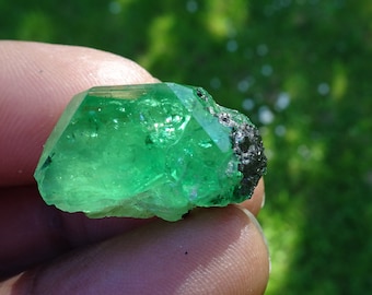 May include: A vibrant green emerald crystal with a rough, natural texture. The crystal has a sharp, geometric shape and is partially covered in a dark gray mineral.