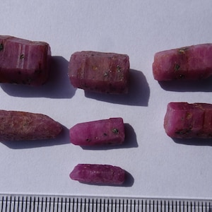 May include: Six rough, pink ruby crystals of varying sizes and shapes. The crystals are on a white surface with a ruler in the background.