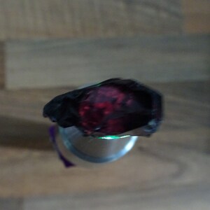 Puede incluir: Primer plano de una piedra preciosa de color rojo oscuro con un brillo verde alrededor de los bordes. La piedra preciosa est&aacute; engastada en un anillo de metal negro.
