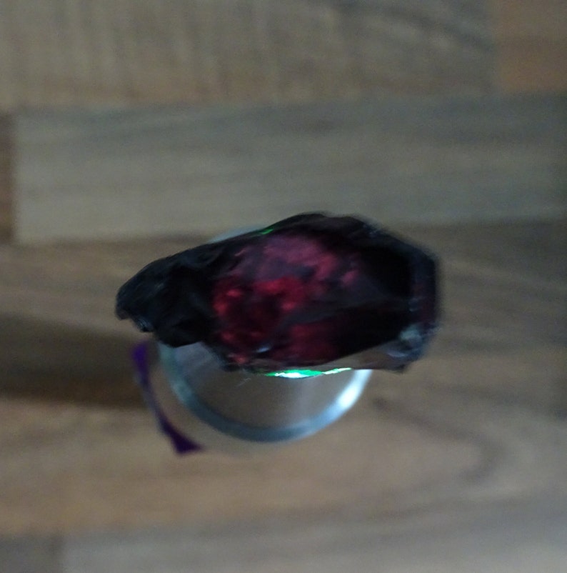 Puede incluir: Primer plano de una piedra preciosa de color rojo oscuro con un brillo verde alrededor de los bordes. La piedra preciosa est&aacute; engastada en un anillo de metal negro.