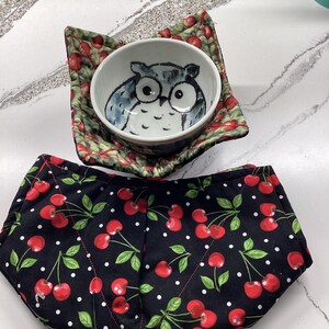 May include: A ceramic bowl with a hand-painted owl design rests on a cherry-patterned bowl cozy. A second bowl cozy with a black background and red cherry pattern is below. The cozy is designed to hold a bowl.