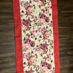 May include: A red table runner with a floral pattern featuring pink and red roses and butterflies. The runner has a green butterfly applique at each end.