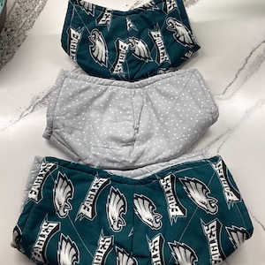 May include: Three bowl cozies. Two are teal with a white and black Eagles logo pattern. The third is gray with a white polka dot pattern. The cozies are quilted and have a curved shape.