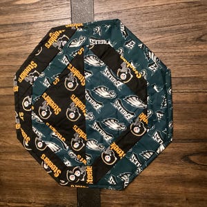 May include: A hexagonal fabric coaster with a patchwork design featuring the Philadelphia Eagles and Pittsburgh Steelers logos. The coaster is made of green, black, and gold fabric.
