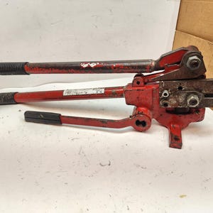 May include: A red metal strapping tool with black handles. The tool has a lever-based design with a cutting mechanism. The handles are textured for grip. The tool appears to be used for securing items with metal straps.