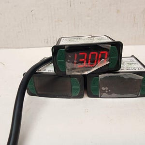 May include: Three black digital temperature controllers with green control buttons on the front panel. The top unit displays the red LED number 13.00. Each device features a rectangular housing with a black power cable attached to the left side.