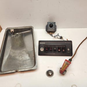 May include: A collection of coffee machine replacement parts including a rectangular stainless steel drip tray, a black control panel with the brand name BUNN and five switches, a small black dial component, a chrome knob, and a red hot water faucet.