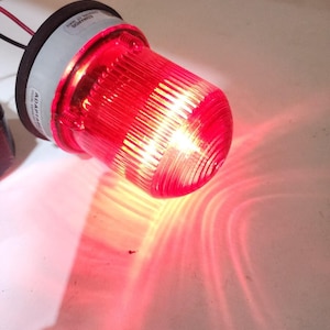 May include: A red emergency light with a ribbed, transparent lens. The light is mounted on a gray and black base with the text "EDWARDS" and "ADAPTA" visible. Wires extend from the base.