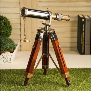 Pu&ograve; includere: Un telescopio in ottone in stile vintage con un treppiede in legno. Il telescopio ha una finitura nera e un tappo dell'obiettivo in ottone. Il treppiede &egrave; in legno scuro e ha tre gambe.