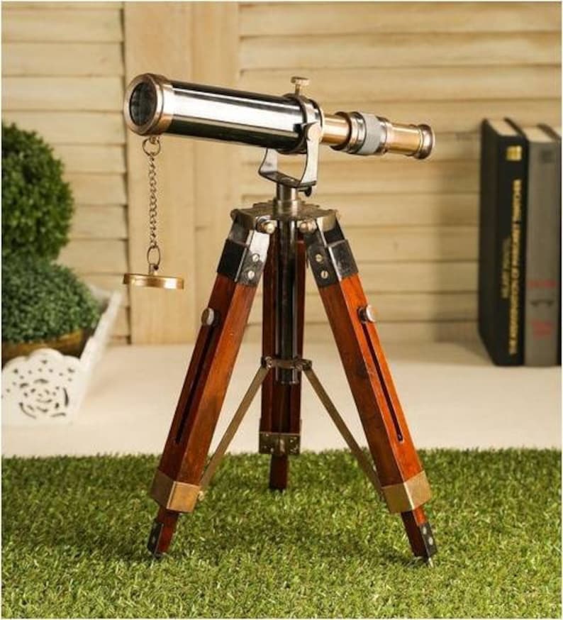 Pu&ograve; includere: Un telescopio in ottone in stile vintage con un treppiede in legno. Il telescopio ha una finitura nera e un tappo dell'obiettivo in ottone. Il treppiede &egrave; in legno scuro e ha tre gambe.