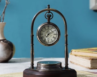 Op de afbeelding: Een messing klok met een standaard, met Romeinse cijfers en de tekst "VICTORIA LONDON 1876". De klok staat op een houten voet en een kaart.