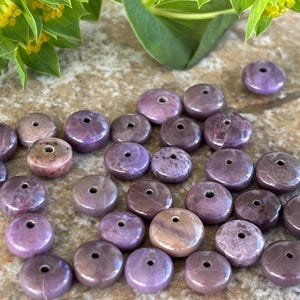 May include: A selection of round, purple gemstone beads, each with a central hole. The beads display varying shades of purple and have a polished surface. Green foliage with yellow flowers is visible in the background.