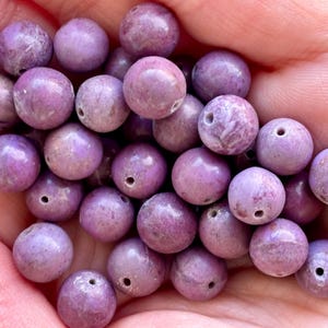 May include: A collection of round, lavender-colored beads with small holes, held in a hand. The beads have a smooth, polished appearance and vary slightly in shade, with some showing subtle marbling. These beads are suitable for jewelry making.