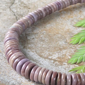 Könnte beinhalten: Eine Halskette aus kleinen, runden, lavendelfarbenen Perlen. Die Perlen sind aneinandergereiht und bilden eine kreisförmige Form. Die Halskette wird auf einer strukturierten, neutral getönten Oberfläche mit grünen Blättern im Hintergrund präsentiert.