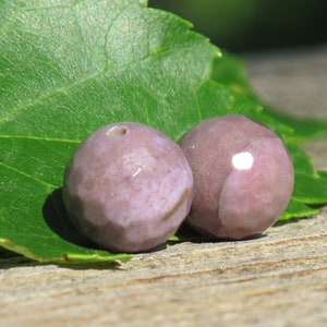 May include: Two faceted, round, lavender-colored beads with a hole drilled through each. The beads are resting on a green leaf, with a blurred wooden background. The beads have a glossy finish and appear to be made of stone.
