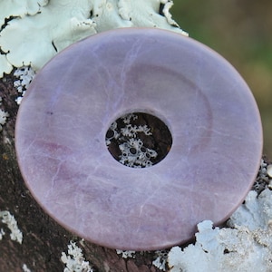May include: A purple stone disc with a hole in the center. The disc is smooth and has a light purple color. It is sitting on a piece of bark with white lichen.