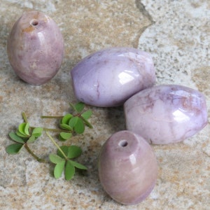 May include: Four smooth, oval-shaped purple and white agate beads. The beads have a polished, glossy finish and are arranged on a light brown textured surface.