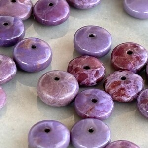 May include: A selection of purple and maroon gemstone beads. The beads are circular with a central hole, and the colours range from pale lavender to deep burgundy. These beads are suitable for jewellery making.