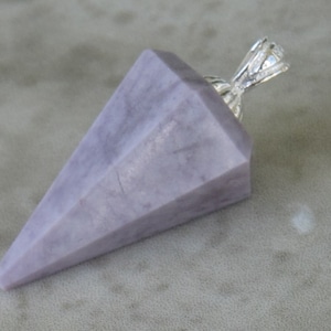 May include: A lavender-colored crystal pendulum with a faceted, conical shape. The pendulum has a silver-colored metal loop at the top for hanging. The crystal is a light purple color.