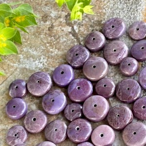 May include: A selection of polished, round, purple gemstones, each with a central drilled hole. The stones range in size and colour, from pale lavender to deep violet. Green foliage is visible in the upper left corner, set against a textured, neutral background.