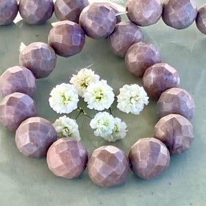 May include: A strand of light purple faceted gemstone beads. The beads are round and have a textured surface. The beads are arranged in a circle and are lying on a light green surface.
