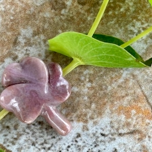 May include: A carved, four-leaf clover in shades of lavender and pink rests on a stem, with a bright green leaf in the background. The clover is a symbol of good luck and is a popular item for jewelry and gifts.